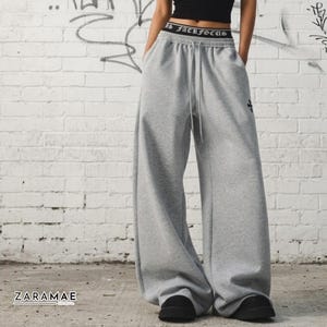 May include: Light gray wide-leg sweatpants featuring a high-rise elastic waistband with the text "JACKFOCUS" in a gothic font. The pants include a drawstring closure, a small black cross emblem on the upper left leg, and a loose, relaxed fit.
