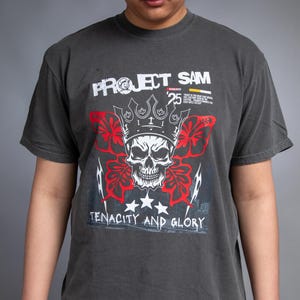 May include: Dark grey t-shirt with a graphic design featuring a skull wearing a crown, red floral accents, and the text "PROJECT SAM" and "TENACITY AND GLORY". The shirt has a crew neck and short sleeves.