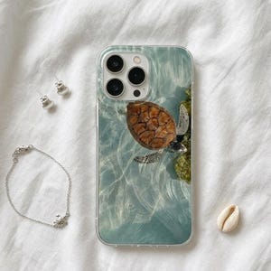 Könnte beinhalten: Eine transparente Handyhülle mit einem Schildkröten-Design, ein silbernes Armband mit einem Schildkröten-Anhänger und silberne Muschelohrringe. Eine weiße Muschel liegt auf einer weißen Stoffoberfläche.