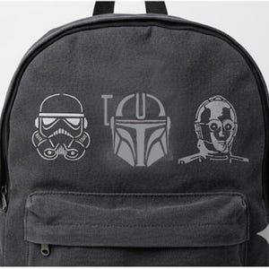 May include: A dark gray backpack featuring three Star Wars character designs in white: a Stormtrooper helmet, the Mandalorian helmet, and C-3PO. The word "I" is between the Stormtrooper and Mandalorian designs. The backpack has a front pocket and a top handle.