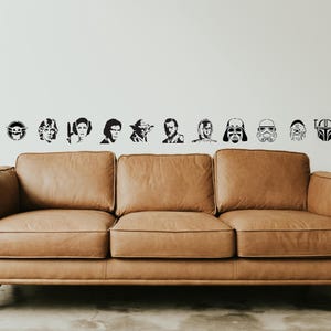 May include: A brown leather sofa with a mid-century modern design. Above the sofa, black and white decals of Star Wars characters and symbols are arranged on a white wall. The sofa has a three-cushion design.