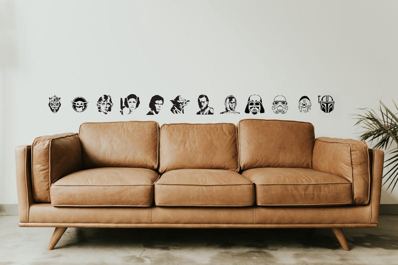 May include: A brown leather sofa with a mid-century modern design. Above the sofa, black and white decals of Star Wars characters and symbols are arranged on a white wall. The sofa has a three-cushion design.