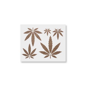 May include: A white square stencil featuring six brown cannabis leaves of varying sizes. The leaves are arranged in a symmetrical pattern, with the largest leaf at the bottom center.