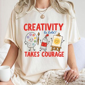 May include: A cream-colored t-shirt featuring a graphic with the red text "CREATIVITY" and "TAKES COURAGE" surrounding illustrations of a smiling paint palette, a paintbrush, a red paint tube, and a sun on a canvas with the text "be bold!"