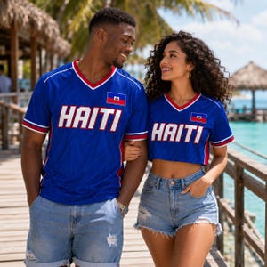 May include: Two blue V-neck shirts featuring the word HAITI in large white letters across the chest and a small Haitian flag emblem. One shirt is a standard length, while the other is a cropped style. Both shirts have red and white trim.