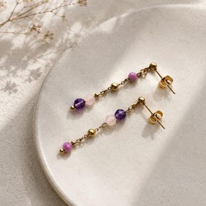 May include: A pair of gold-colored dangle earrings. Each earring features a delicate chain with small gold beads and a series of colorful gemstones. The gemstones include purple, pink, and light pink beads. The earrings are displayed on a white plate.