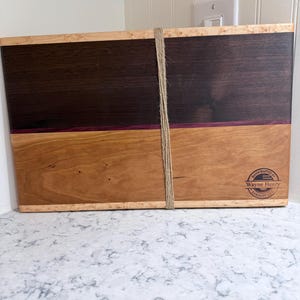 May include: A handcrafted wooden cutting board with a two-tone design. The top half is dark brown, the bottom half is light brown, and a thin purple line separates them. A natural twine is wrapped around the board. The board has a logo that says "Handcrafted by Wayne Henry Sawdust Lab".