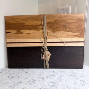May include: A rectangular wooden cutting board with a two-tone design. The top section is light brown with natural wood grain, the middle is a thin strip of dark red, and the bottom is dark brown. The board is tied with twine and has a small tag.