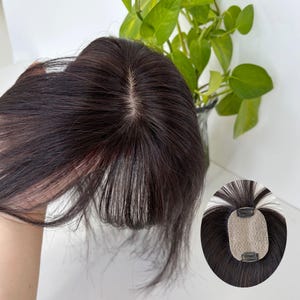 May include: Dark brown hair topper with a natural-looking part. The hairpiece has a clip-in design for easy attachment. A small, close-up image shows the clip-in mechanism and the blend of dark and light strands.