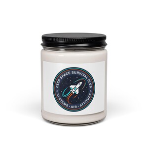 May include: A white candle jar with a black lid. The jar has a label with a circular design featuring a rocket ship and the text "DEEP SPACE SURVIVAL CLUB SYSTEMS AIR ATTITUDE". The candle is a home decor item.