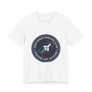 May include: White t-shirt featuring a circular graphic with a rocket ship design. The graphic includes the text "DEEP SPACE SURVIVAL CLUB" and other text. The design is in shades of blue, orange, and white.
