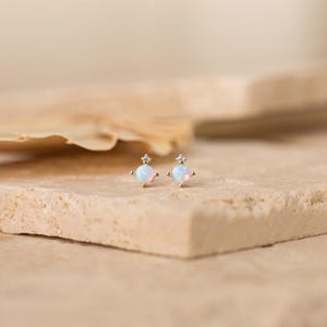 May include: A pair of gold stud earrings featuring a round, iridescent opal stone set in a four-prong setting, topped with a single, tiny, clear, round-cut gemstone. The earrings are crafted from a polished gold-tone metal with a delicate design.
