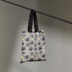 May include: A grey tote bag featuring a repeating pattern of small black fuzzy circular creatures with large white eyes, interspersed with tiny yellow and light pink star-shaped icons. The bag has two black fabric handles attached at the top.