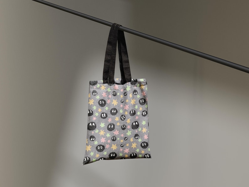 May include: A grey tote bag featuring a repeating pattern of small black fuzzy circular creatures with large white eyes, interspersed with tiny yellow and light pink star-shaped icons. The bag has two black fabric handles attached at the top.