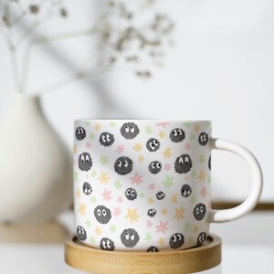 May include: A white ceramic mug featuring a repeating pattern of small, fuzzy, dark gray soot sprites with large white eyes, interspersed with scattered pastel pink, yellow, and light green star shapes across the entire exterior surface.