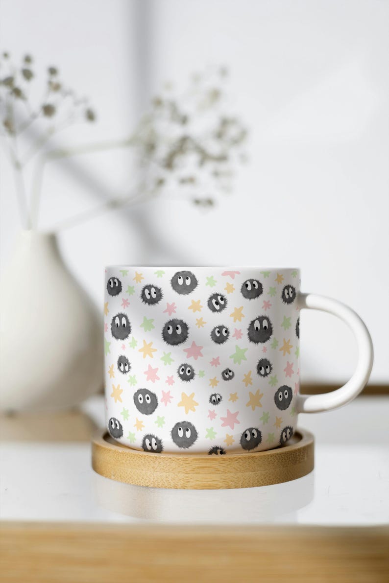 May include: A white ceramic mug featuring a repeating pattern of small, fuzzy, dark gray soot sprites with large white eyes, interspersed with scattered pastel pink, yellow, and light green star shapes across the entire exterior surface.