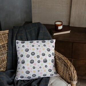 May include: A square throw pillow with a white background featuring a repeating pattern of small, dark gray, fuzzy circular creatures with large white eyes, interspersed with light pink, yellow, and pale green star-shaped accents.