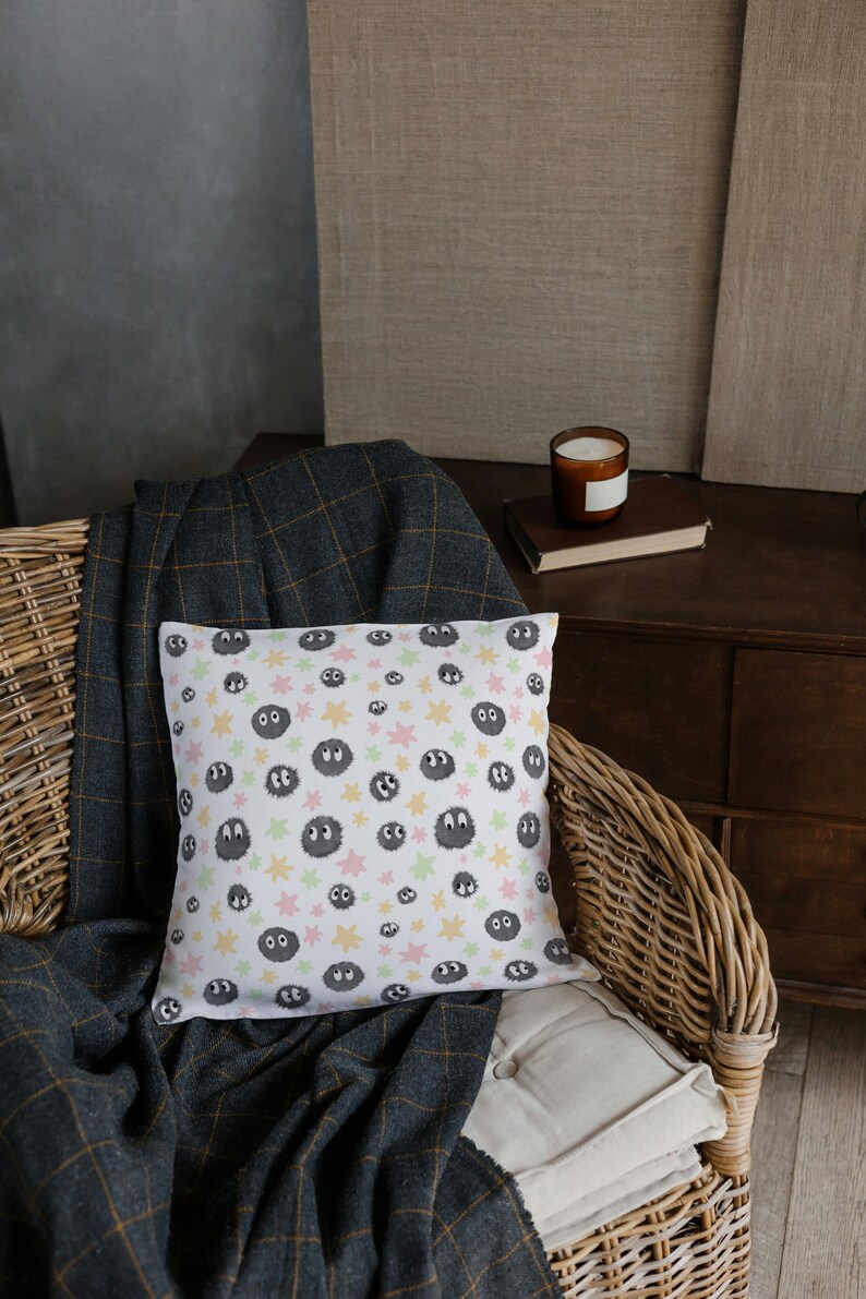 May include: A square throw pillow with a white background featuring a repeating pattern of small, dark gray, fuzzy circular creatures with large white eyes, interspersed with light pink, yellow, and pale green star-shaped accents.