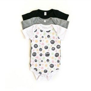 May include: A stack of three short-sleeve baby bodysuits. The top bodysuit is white with a repeating pattern of dark gray fuzzy circular creatures and small pastel-colored stars. Beneath it are a heather gray bodysuit and a solid black bodysuit.
