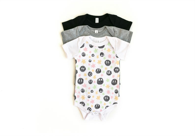 May include: A stack of three short-sleeve baby bodysuits. The top bodysuit is white with a repeating pattern of dark gray fuzzy circular creatures and small pastel-colored stars. Beneath it are a heather gray bodysuit and a solid black bodysuit.