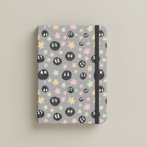 May include: A gray notebook featuring a repeating pattern of small black fuzzy circular creatures with white eyes, interspersed with pastel pink, yellow, and light green star shapes. The notebook includes a vertical black elastic closure band.