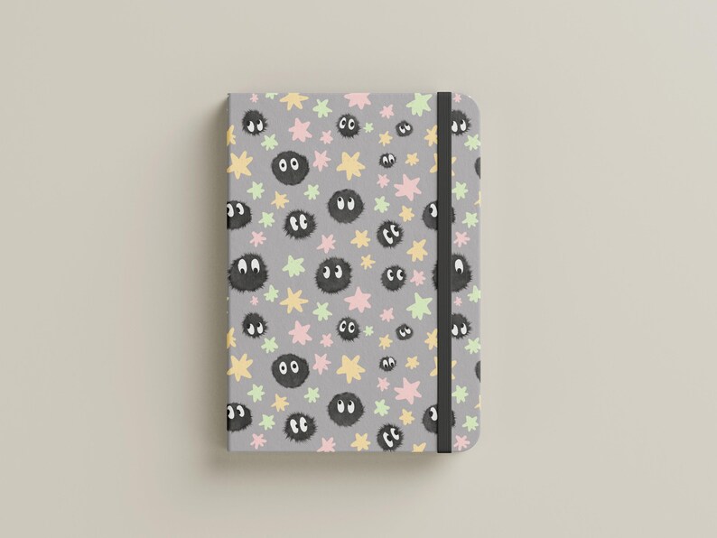 May include: A gray notebook featuring a repeating pattern of small black fuzzy circular creatures with white eyes, interspersed with pastel pink, yellow, and light green star shapes. The notebook includes a vertical black elastic closure band.
