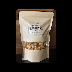 Könnte beinhalten: Ein brauner Papiersack mit Maple Puffcorn und einem transparenten Fenster. Der Beutel hat ein Etikett mit dem Text "Tait's Flat Rock Maple Farm" und "Maple Puffcorn". Die Zutaten sind unten aufgeführt. Der Beutel ist versiegelt.