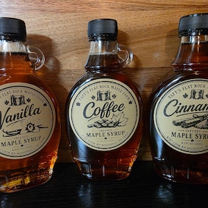 May include: Three glass bottles of Taft's Flat Rock Maple Farm infused maple syrup. The bottles contain vanilla, coffee, and cinnamon flavours, each with a black cap, a handle, and a circular label displaying the flavour name and 250ml volume.