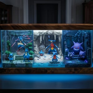 Puede incluir: Una vitrina decorativa e iluminada con tres escenas distintas de Pokémon. Las escenas incluyen un paisaje de piedra, una cueva y un entorno gótico, todo dentro de una caja de vidrio con marco de madera. La base emite un brillo azul.