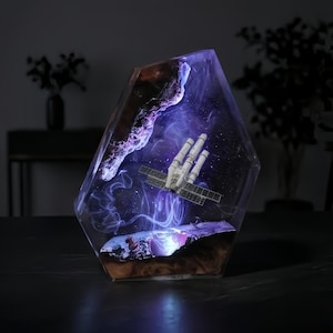 May include: A decorative resin sculpture featuring a space station model encased within a clear, geometric form. The interior showcases a miniature galaxy with swirling purple and white hues, and brown and black accents.