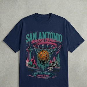 Pu&ograve; includere: T-shirt blu navy con grafica di basket. Il design include il testo "SAN ANTONIO BASKETBALL", un pallone da basket in una rete, uno skyline e il testo "BUILT DIFFERENT DRIVEN TO WIN".