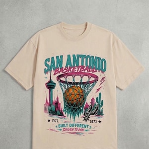 Pu&ograve; includere: T-shirt beige con grafica a tema basket. Il design include le parole "San Antonio Basketball" sopra un pallone da basket in una rete. Sotto ci sono le parole "Built Different, Driven to Win".