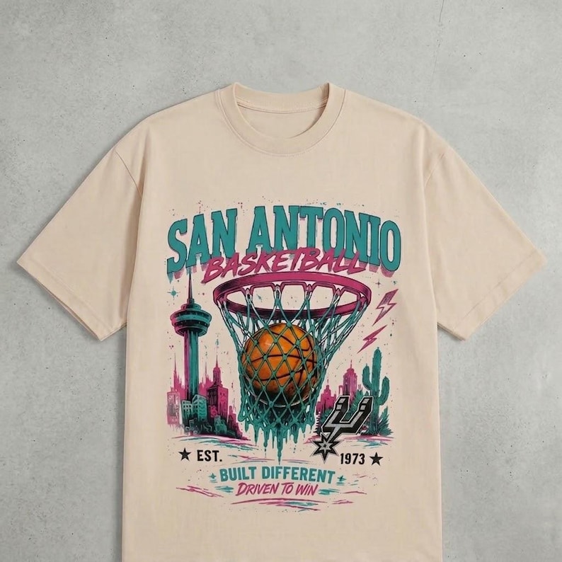 Pu&ograve; includere: T-shirt beige con grafica a tema basket. Il design include le parole "San Antonio Basketball" sopra un pallone da basket in una rete. Sotto ci sono le parole "Built Different, Driven to Win".