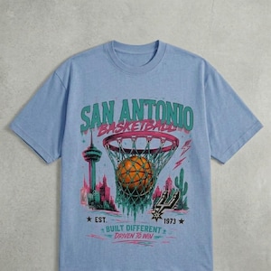 Pu&ograve; includere: T-shirt azzurra con grafica di basket, rete e skyline. Il testo recita "SAN ANTONIO BASKETBALL", con "EST. 1973" e "BUILT DIFFERENT DRIVEN TO WIN". Il design &egrave; nei toni del verde, rosa e arancione.