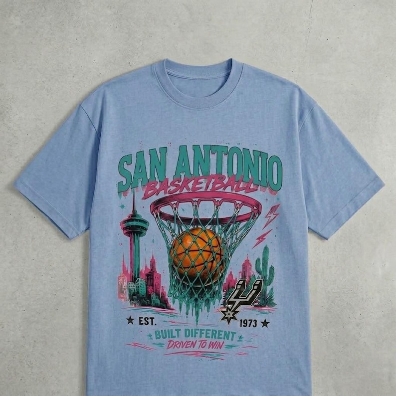 Pu&ograve; includere: T-shirt azzurra con grafica di basket, rete e skyline. Il testo recita "SAN ANTONIO BASKETBALL", con "EST. 1973" e "BUILT DIFFERENT DRIVEN TO WIN". Il design &egrave; nei toni del verde, rosa e arancione.