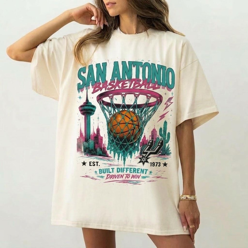 Pu&ograve; includere: T-shirt oversize color crema con grafica di basket di San Antonio. Il design include un pallone da basket che entra in una rete, un paesaggio urbano e il testo "SAN ANTONIO BASKETBALL" in verde acqua e rosa. La maglietta ha anche il testo "EST. 1973".