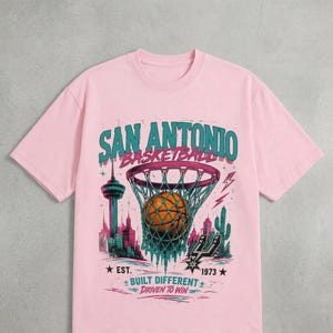 Pu&ograve; includere: T-shirt rosa chiaro con grafica di basket di San Antonio. Il design presenta un pallone da basket in una rete, il testo "SAN ANTONIO BASKETBALL" e il testo "BUILT DIFFERENT DRIVEN TO WIN".