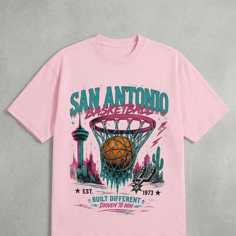 Pu&ograve; includere: T-shirt rosa chiaro con grafica di basket di San Antonio. Il design presenta un pallone da basket in una rete, il testo "SAN ANTONIO BASKETBALL" e il testo "BUILT DIFFERENT DRIVEN TO WIN".