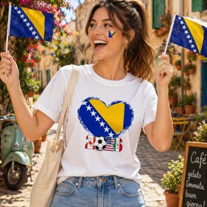 May include: A white t-shirt featuring a heart-shaped design with the flag of Bosnia and Herzegovina, a soccer ball, and the number 2026. The person is holding two small flags of Bosnia and Herzegovina.