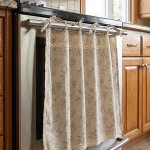 May include: A beige kitchen towel with a floral pattern and lace trim, hanging from a stainless steel oven. The towel features a delicate design of small pink and blue flowers and green leaves.