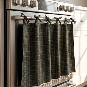 Puede incluir: Una cortina de horno a cuadros verde oscuro y dorado con ribete de flecos blancos, atada a la manija del horno con lazos verdes oscuros. La cortina cuelga de un horno de acero inoxidable.