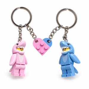 May include: Two keychains featuring plastic minifigures dressed in shark costumes, one pink and one blue. Each figure is attached to a metal ring and chain, connected by a central heart-shaped plastic brick piece split into pink and blue halves.