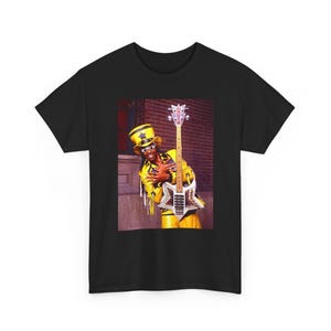 May include: A black t-shirt featuring a graphic print of a person wearing a yellow suit and a matching top hat, holding a star-shaped electric bass guitar. The person wears star-shaped sunglasses and the guitar headstock displays the text Bootsy.