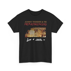 Peut inclure: Un t-shirt noir à manches courtes avec une impression graphique de Johnny Thunders & The Heartbreakers. Le motif comprend le texte "LIVE AT MAX'S KANSAS CITY '79" et une photographie de concert sépia sur le devant du vêtement.