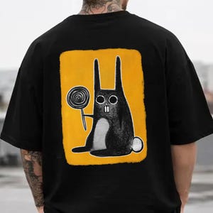 May include: Black t-shirt featuring a cartoon rabbit holding a lollipop on a yellow background. The rabbit is black with a white belly and a white tail. The t-shirt has short sleeves and a crew neck.
