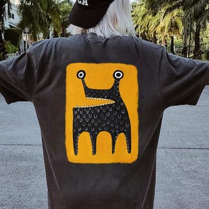 May include: Dark gray t-shirt with a yellow rectangle on the back featuring a black creature illustration. The creature has two eyes, a mouth with teeth, and three legs. The shirt is a casual style.