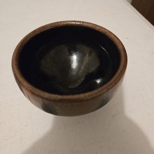 May include: A small, round ceramic bowl with a smooth, glossy black interior and a textured, matte brown exterior. The rim of the bowl is thick and rounded, featuring a mottled brown and tan finish that contrasts with the dark, reflective center.