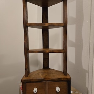 May include: A brown wooden corner shelf unit featuring three open triangular tiers and a base section with two small drawers. Each drawer is equipped with a single round white knob. The unit has curved side supports and a dark wood finish.