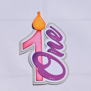 Könnte beinhalten: Ein mehrschichtiger Papierauschnitt in Form einer Kerze mit der Zahl "1" und dem Wort "One". Die Kerzenflamme ist orange, die Zahl ist rosa und das Wort "One" ist lila. Der Ausschnitt hat einen grauen Rand.