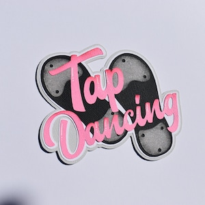 May include: A layered paper cutout featuring two tap shoes in black and gray, with the words "Tap Dancing" in pink script. The design has a white and gray border, set against a white background.